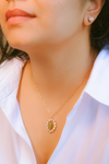 Close-up of a person wearing a gold necklace with a green pendant and white stones, against a blurred background.