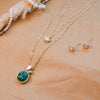 Gold necklace with green pendant and matching earrings on a beige surface